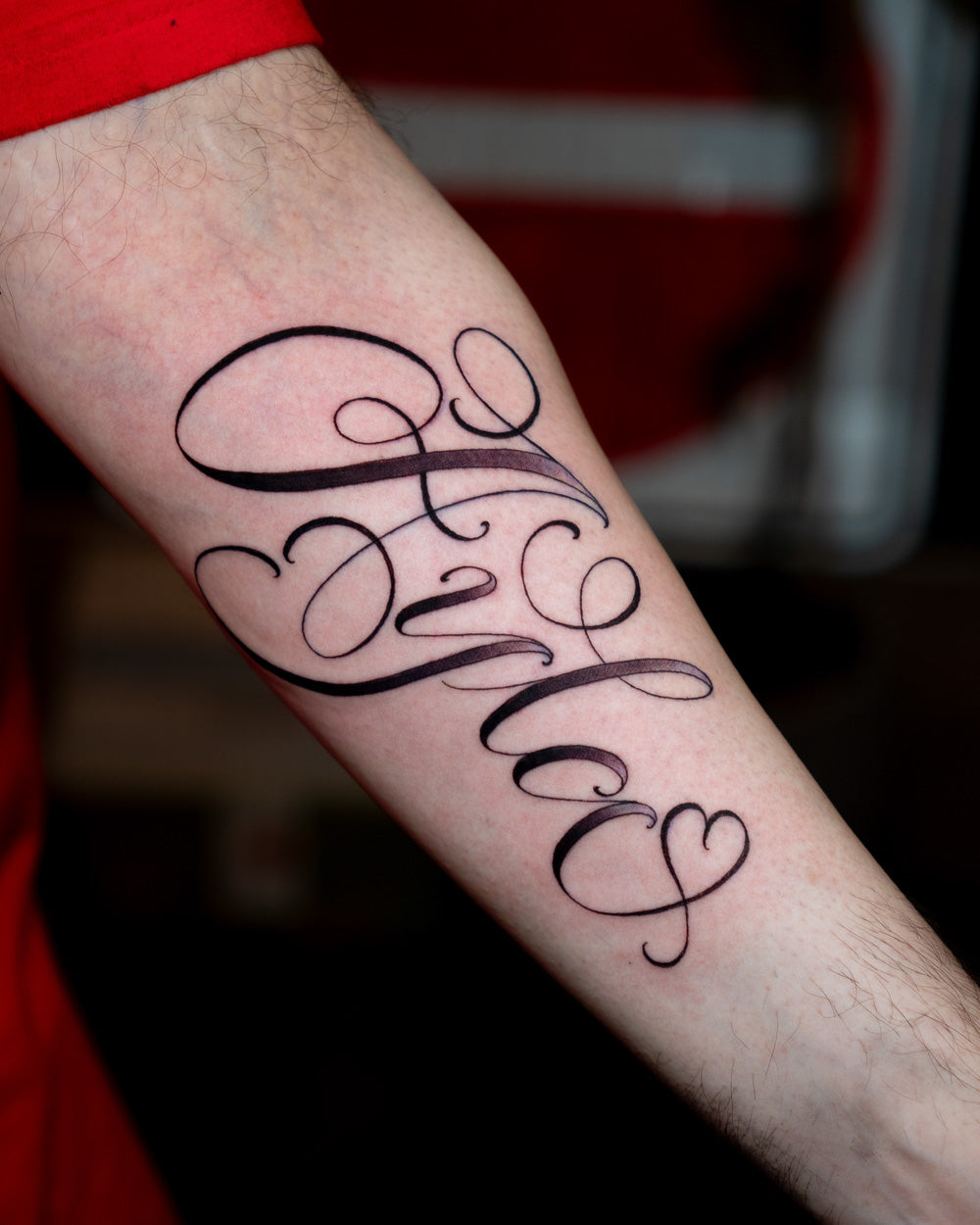 Black ink script lettering name tattoo reading “Ayla” with a small heart on the forearm