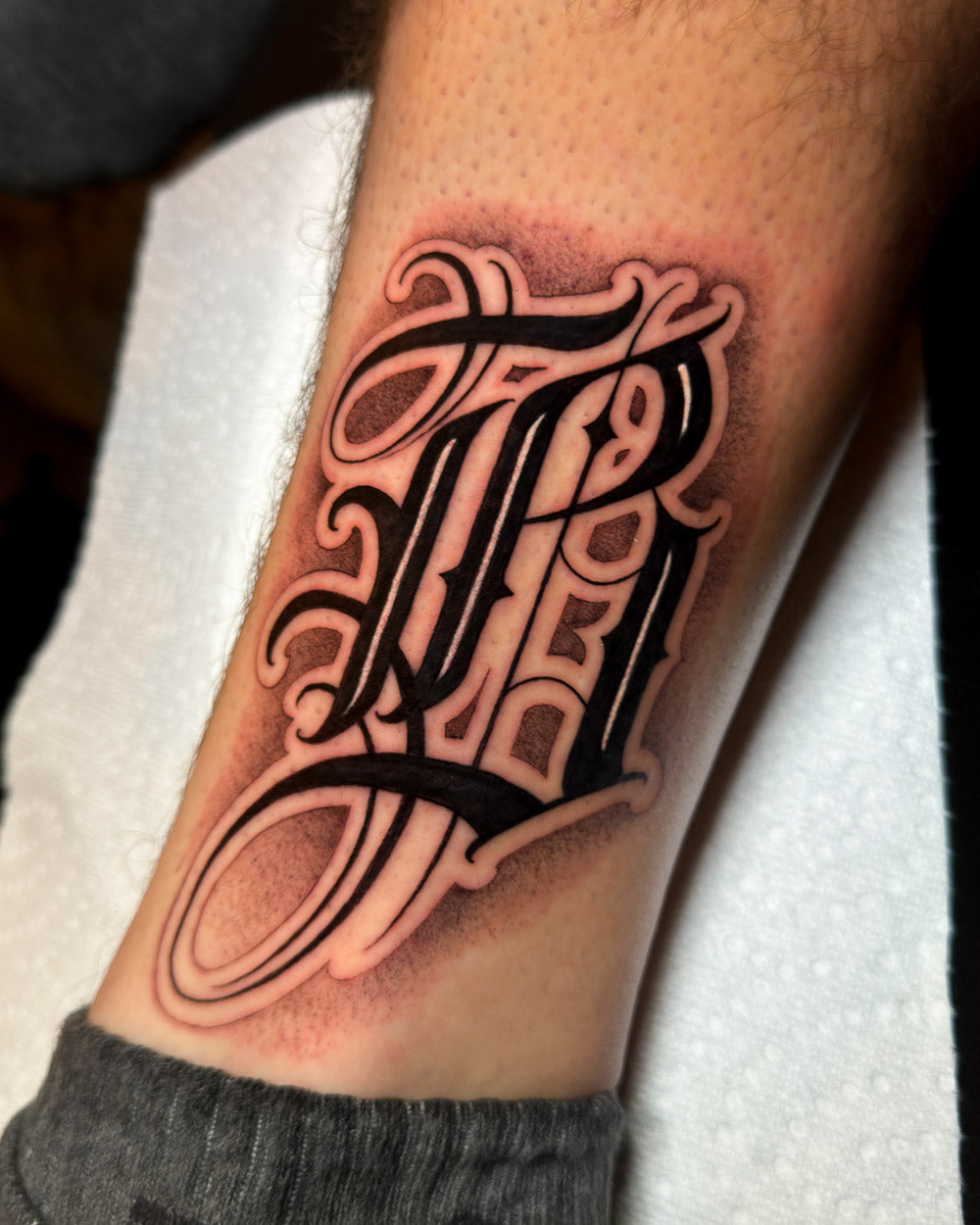 Black and grey Chicano script lettering tattoo with bold flowing calligraphic forms on the forearm