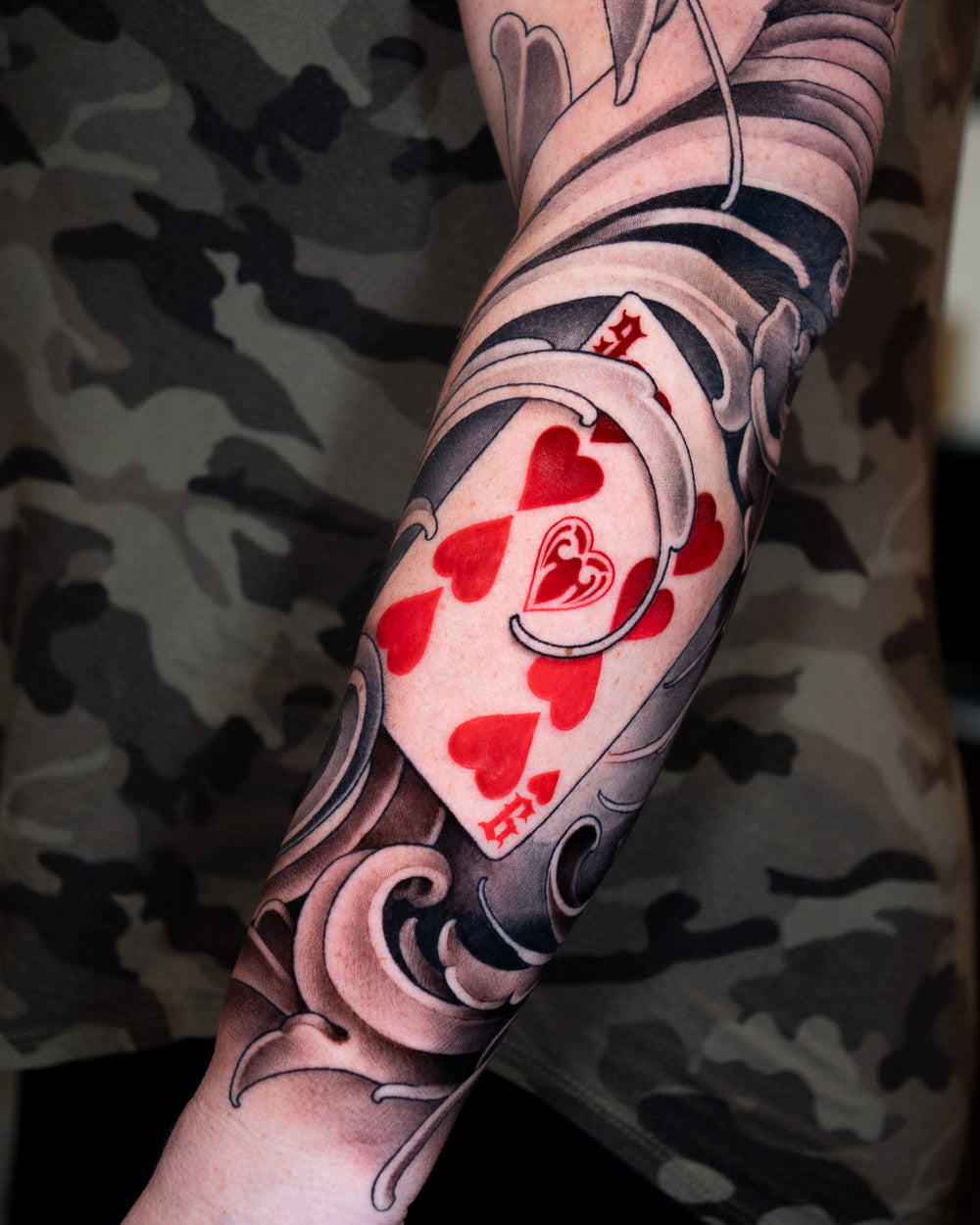 Nine of hearts playing card tattoo with red hearts surrounded by black and grey filigree on the forearm