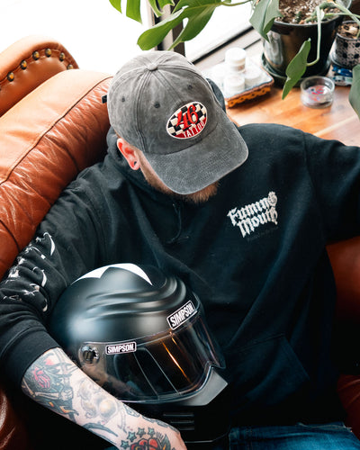 Person wearing a black sweatshirt, a 46 tattoo branded cap, holding a Simpson helmet, sitting on a brown leather couch.