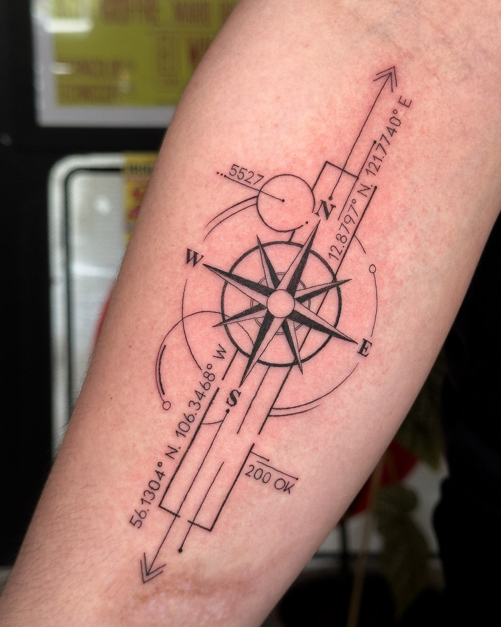 Black and grey linework compass tattoo with geographic coordinates and directional markings on the forearm